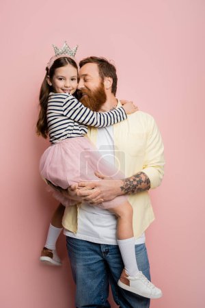 Tätowierter Mann hält fröhliche Tochter mit Krone-Haarreif auf rosa Hintergrund 