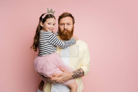 Lächelndes Mädchen im Kronenstirnband mit Hasenohren-Geste in der Nähe des Kopfes eines ernsten Vaters auf rosa Hintergrund 
