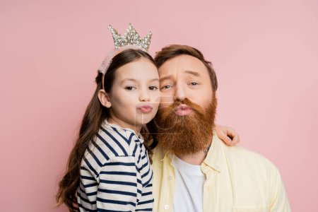 Preteen daughter with crown headband pouting lips and hugging dad isolated on pink  