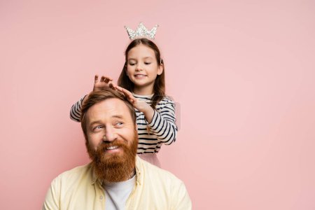 Fröhliches Mädchen mit Krone Stirnband berührt Haar des lächelnden Papas isoliert auf rosa  
