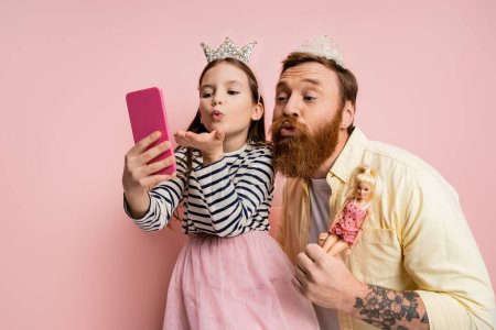 Foto de Hija y padre con muñecas y diademas de corona tomando selfie en el teléfono inteligente sobre fondo rosa - Imagen libre de derechos