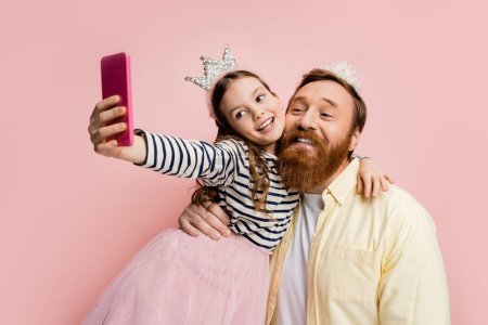 Foto de Sonriente chica abrazando padre en diadema de corona y tomando selfie aislado en rosa - Imagen libre de derechos