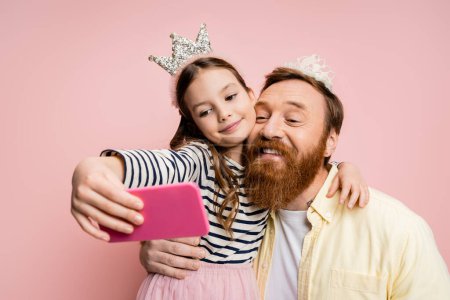 Foto de Hombre positivo con diadema de corona tomando selfie con hija aislada en rosa - Imagen libre de derechos