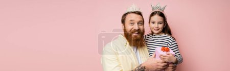 Positive man and daughter with crown headbands holding gift isolated on pink, banner 