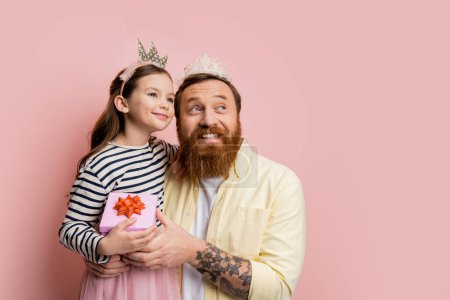 Smiling daughter and dad with crown headbands holding gift box on pink background 