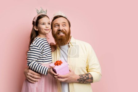 Positive father and daughter with crown headbands holding present and looking away on pink background 