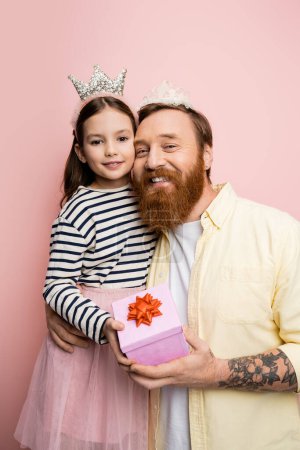 Cheerful man and child in crown headbands holding present on pink background 