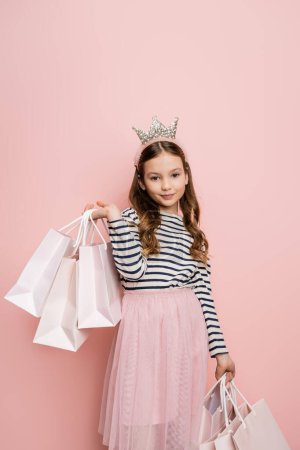 Photo pour Enfant préadolescent insouciant dans le bandeau de la couronne tenant des sacs à provisions sur fond rose - image libre de droit