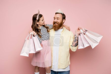 Photo pour Fille gaie tenant des sacs à provisions et câlin père avec couronne bandeau sur fond rose - image libre de droit
