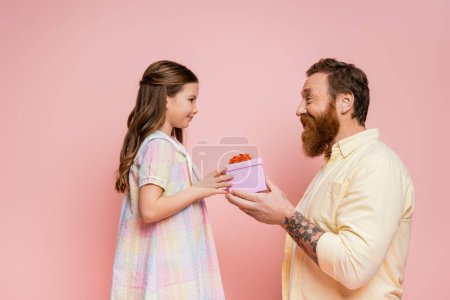 Téléchargez les photos : Vue latérale de l'homme barbu joyeux donnant cadeau à la fille isolée sur rose - en image libre de droit