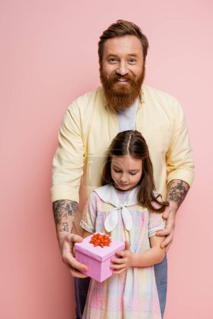Carefree man giving gift box to preteen daughter on pink background 