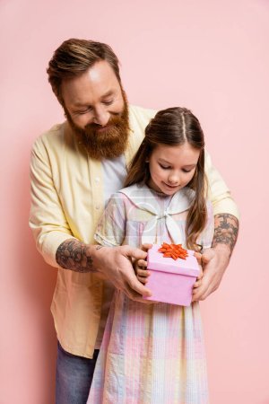 Tattooed man holding gift near preteen daughter on pink background 