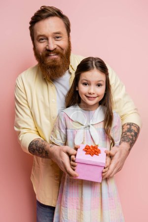 Positive tattooed man holding gift box near daughter on pink background 