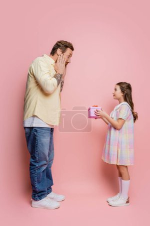 Téléchargez les photos : Vue latérale de l'homme choqué regardant la fille préadolescente avec un cadeau sur fond rose - en image libre de droit