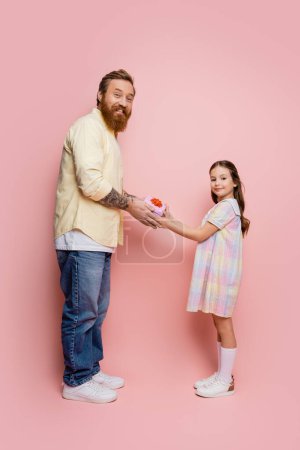 Chica positiva dando regalo a papá barbudo y mirando a la cámara en el fondo rosa 