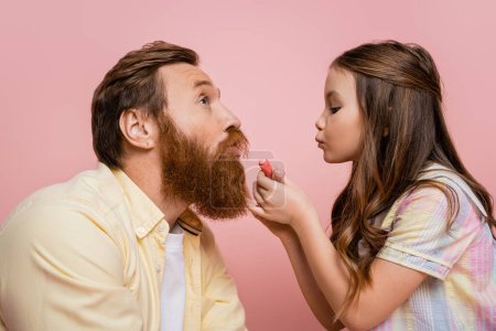 Girl holding lipstick and pouting lips near bearded father isolated on pink  