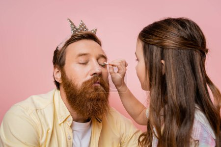 Preteen girl applying eyeshadow on father with crown headband isolated on pink  