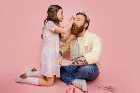 Foto de Chica preadolescente aplicando cosmética decorativa en papá tatuado con diadema de corona sobre fondo rosa - Imagen libre de derechos