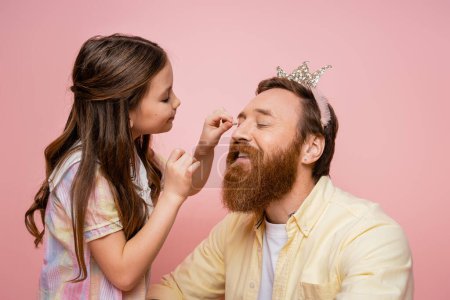 Smiling preteen girl applying eyeshadow on bearded dad with crown headband isolated on pink  