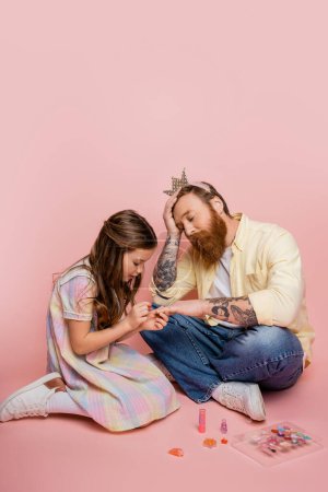 Photo for Preteen kid applying nail polish on hand of upset dad with crown headband on pink background - Royalty Free Image