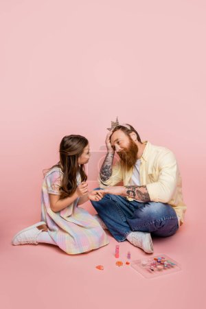 Preteen girl holding nail lack near hand of tired father with crown headband on pink background 