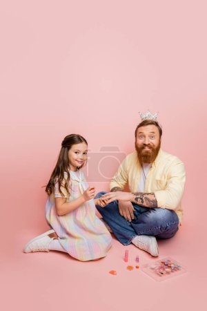 Cheerful girl holding nail polish near father with crown headband and decorative cosmetic on pink background 