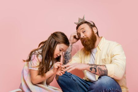Preteen girl applying nail polish on hand of tattooed dad with crown headband on pink background 