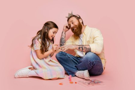 Photo for Cheerful girl applying nail polish on hand of tattooed dad with crown headband on pink background - Royalty Free Image