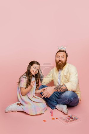Positive girl holding nail polish near tattooed dad with crown on head and decorative cosmetics on pink background 