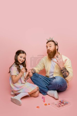 Sonriente niño sosteniendo esmalte de uñas cerca de padre con corona en la cabeza haciendo pucheros labios sobre fondo rosa 