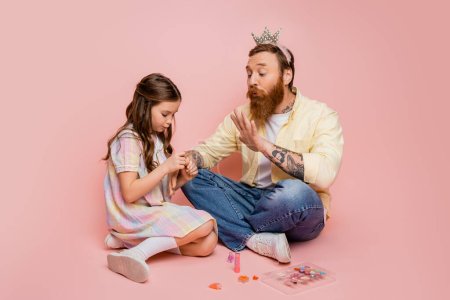 Man with crown on head blowing on hand near daughter with nail polish on pink background 