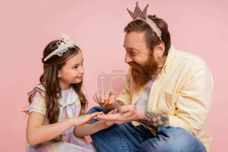 Hombre alegre con corona en la cabeza sosteniendo esmalte de uñas cerca de hija preadolescente sobre fondo rosa 