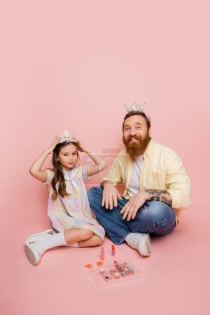 Positive bearded man looking at camera near daughter with crown and decorative cosmetics on pink background 