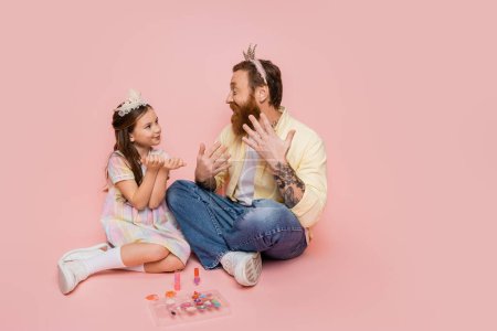 Fröhlicher Vater und Tochter mit Kronen-Stirnbändern sitzen neben dekorativer Kosmetik auf rosa Hintergrund 