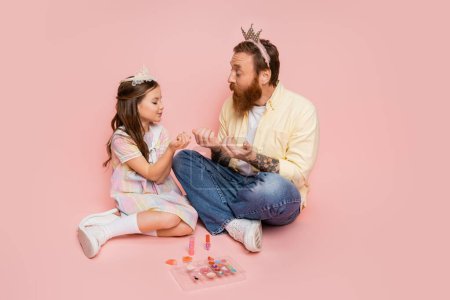 Bearded man with crown headband looking at daughter near decorative cosmetics on pink background 