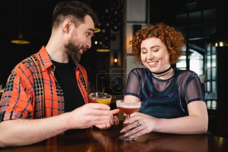Positive man holding negroni cocktail and talking to redhead friend in bar 