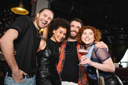 Positive redhead woman holding foam cocktail near interracial friends looking at camera and hugging in bar 