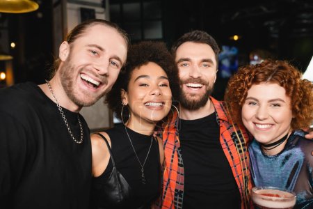 Smiling curly woman holding cocktail near multiethnic friends looking at camera while spending time in bar 