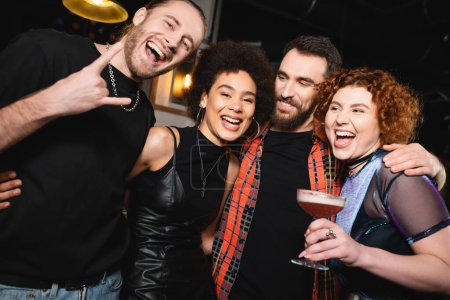 Aufgeregte Frau hält Klee-Club-Cocktail in der Nähe unbeschwerter interrassischer Freunde und blickt in Bar in die Kamera 