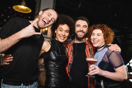 Fröhliche rothaarige Frau hält Cocktail in der Nähe multiethnischer Freunde und blickt in Bar in die Kamera 
