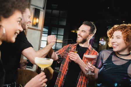 Positive men holding hands near happy interracial friends with cocktails in bar 