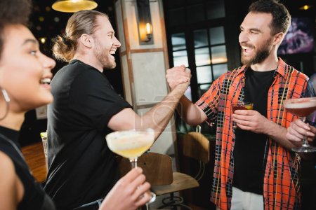 Cheerful bearded friends with cocktails holding hands while meeting in bar 