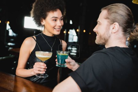 Sonriente mujer afroamericana sosteniendo pisco sour cocktail cerca borrosa amigo en bar 