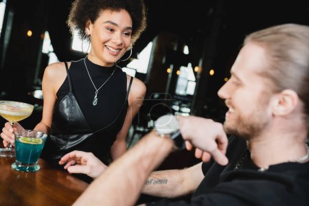 Enthousiaste femme afro-américaine tenant pisco cocktail aigre près d'ami flou dans le bar 