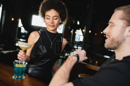 Joven afroamericana mujer tomando pisco sour cocktail desde stand cerca borrosa amigo en bar 
