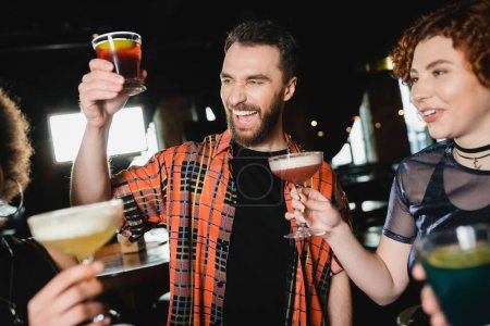 Fröhlicher bärtiger Mann hält Negroni-Cocktail in der Nähe von Freunden mit Gläsern in Bar 