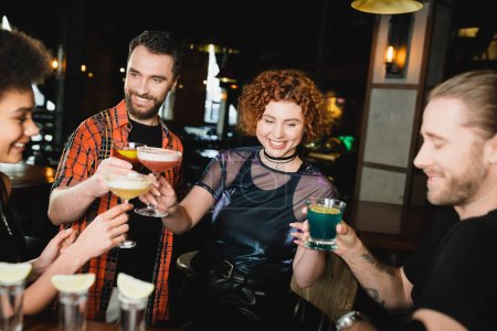 Mujer pelirroja sonriente tintineando cócteles con amigos multiétnicos en el bar 