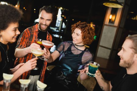 Overjoyed multiethnic friends holding glasses with cocktails near blurred tequila shots in bar 
