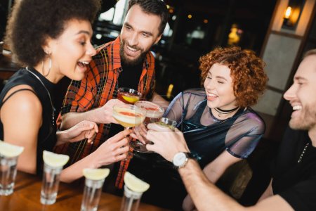 Gente alegre multiétnica tintineando diferentes cócteles cerca de tragos de tequila en el bar 