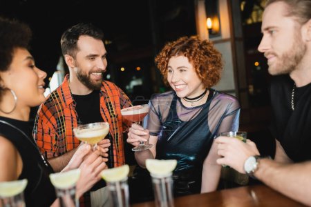 Mujer alegre sosteniendo cóctel cerca de amigos multiétnicos y tequila disparos en el bar 